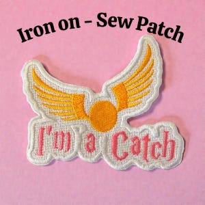 May include: Embroidered patch with a white border, featuring gold wings and a central orange circle. The text "I'm a Catch" is in pink, with the words "Iron on - Sew Patch" above. The patch is set against a pink background.