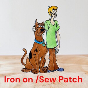 May include: Embroidered patch featuring Scooby-Doo and Shaggy from the cartoon. Scooby-Doo is brown with black spots and a blue collar. Shaggy wears a green shirt and brown trousers. The text "Iron on / Sew Patch" is at the bottom.