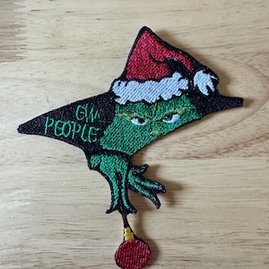 May include: Embroidered patch featuring a green character wearing a red Santa hat with white trim. The character's face has an angry expression. The patch includes the text "GM PEOPLE" in green. A red ornament hangs from the character's hand.