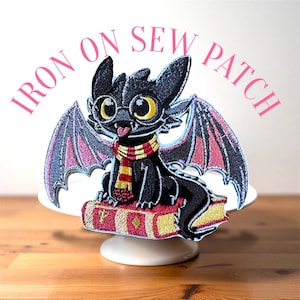 Peut inclure: Écusson thermocollant brodé représentant un dragon noir avec de grands yeux jaunes, des lunettes et une écharpe rouge et or. Le dragon est assis sur un livre avec des lettres dorées. Le patch porte l'inscription "IRON ON SEW PATCH" en rose.