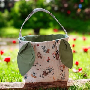 May include: A fabric Easter basket with a handle and green accents. The basket features a cream-colored background with a pattern of woodland animals and floral designs. The basket has two green, leaf-shaped side accents.