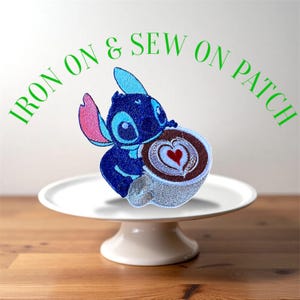 May include: Embroidered patch featuring the character Stitch from Lilo & Stitch, holding a coffee cup with a heart design. The patch is blue, pink, and brown. Text above the patch reads "IRON ON & SEW ON PATCH". The patch is displayed on a white cake stand.