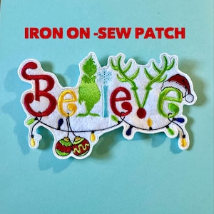 May include: A white felt patch with the word "Believe" in red, yellow, and green, decorated with Christmas lights, ornaments, and a Santa hat. The text "IRON ON -SEW PATCH" is above the word "Believe".