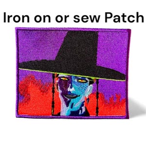 Puede incluir: Parche bordado con un rostro estilizado con sombrero negro sobre fondo morado. El rostro tiene piel azul y morada, ojos amarillos y detalles rojos. El texto "Iron on or sew Patch" está en la parte superior.