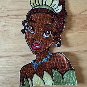 May include: Embroidered patch featuring a princess with brown skin, brown hair, and a green and yellow crown. She wears a blue necklace and a green dress. The patch is on a wooden surface.