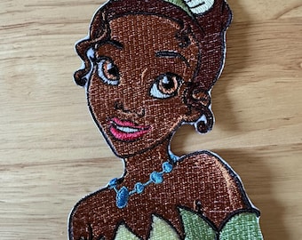 Op de afbeelding: Geborduurde patch met een prinses met bruine huid, bruin haar en een groene en gele kroon. Ze draagt een blauwe ketting en een groene jurk. De patch ligt op een houten oppervlak.