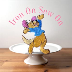 May include: Embroidered patch featuring a cartoon kangaroo in a blue shirt and pink ears, with the text "Iron On Sew On" in pink. The patch is displayed on a white cake stand against a light background and wooden surface.