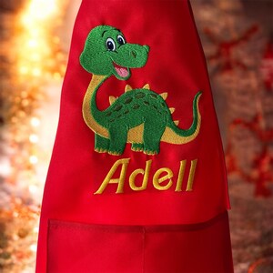 May include: Red apron with a green dinosaur embroidery and the name "Adell" in yellow. The dinosaur has a yellow neck and belly, with green spots on its back. The apron has a pocket.