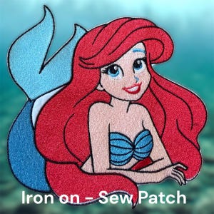 May include: Embroidered patch featuring a cartoon mermaid with red hair, blue eyes, and a blue seashell top. The mermaid has a blue tail and is smiling. The text "Iron on - Sew Patch" is at the bottom.
