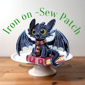 Peut inclure: Écusson brodé représentant un dragon noir aux grands yeux jaunes, portant une écharpe rouge et dorée, posé sur une pile de livres. L'écusson porte les mots "Iron on - Sew Patch" en vert.