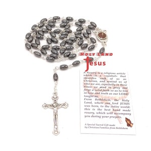 May include: A rosary with dark gray oval beads and a silver crucifix. The rosary is made of linked beads and a silver chain. A small card with text is visible in the image.