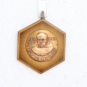 May include: A hexagonal wooden plaque with a gold-colored relief of a man's face. The plaque has the text "Servus Dei Valentinus" around the image.