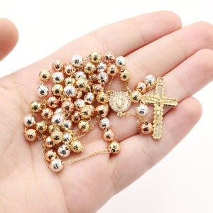 Könnte beinhalten: Eine goldene Rosenkranz Halskette mit einem Kreuzanhänger. Die Perlen sind aus drei verschiedenen Goldfarben gefertigt: Gelb, Weiß und Rosé.