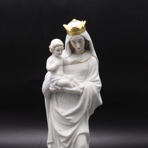 Può includere: Statua in porcellana bianca della Vergine Maria che tiene in braccio un bambino Gesù. Maria indossa una corona e una lunga veste. La statua è su una base bianca.