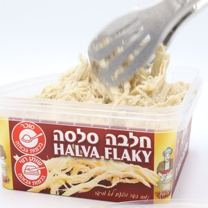 May include: A clear plastic container filled with a white, stringy halva. The container has a yellow label with the text "Halva Flaky" in Hebrew and English. A silver tongs is lifting some of the halva out of the container.
