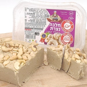 May include: A block of halva, a Middle Eastern confection, topped with cashews, sits on a wooden cutting board. The halva is light brown and has been sliced into pieces. A clear plastic container with a label is in the background.