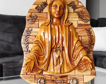 May include: A wooden wall plaque featuring a carved image of the Virgin Mary with the words "God Bless Our Home" on the top. The plaque is made of olive wood and has a natural wood finish. It is decorated with a wreath of olive branches and the word "Bethlehem" is inscribed on the bottom.