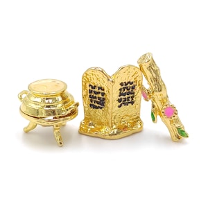 May include: A set of three gold-toned miniature religious objects. The objects are a pot with a lid, two stone tablets with Hebrew text, and a branch with pink flowers and green leaves.