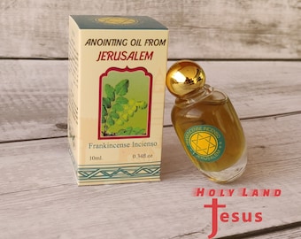 May include: A small glass bottle of anointing oil from Jerusalem, labeled "Frankincense Incienso" with a green and gold label. The bottle is sitting on a wooden surface with a box behind it. The box is labeled "Anointing Oil from Jerusalem" and has a picture of a green plant on it. The box also says "Frankincense Incienso" and has the measurements "10ml. 0.34fl.oz".