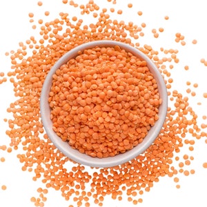 May include: A white bowl filled with red lentils, surrounded by scattered red lentils on a white background.