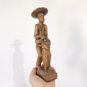 Puede incluir: Figura de madera tallada de una persona con sombrero que sostiene un objeto cilíndrico. La figura es de color marrón claro y se apoya sobre una base rectangular. El sombrero es de ala ancha.