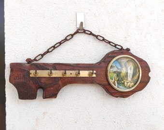 Könnte beinhalten: Ein schlüsselförmiger Schlüsselhalter aus Holz mit fünf Haken und einem runden Bild der Jungfrau Maria in Lourdes, Frankreich. Der Halter hängt an einer rostigen Kette.