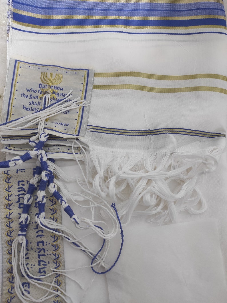 May include: A white prayer shawl with blue and gold stripes. The shawl has fringes and a small tag with a quote in Hebrew and English. The tag reads "But to you who fear My name the Sun of righteousness shall shine on you and healing in its wings."