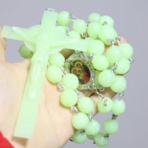 Może przedstawiać: Zielony różaniec świecący w ciemności z srebrnym krucyfiksem i srebrnym medalem z religijnym wizerunkiem. Korale różańca są wykonane z plastiku i nawleczone na srebrny łańcuszek.