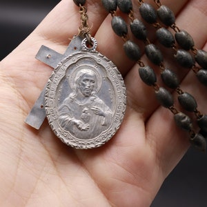 Puede incluir: Un rosario antiguo con cuentas oscuras de forma ovalada y un colgante plateado. El colgante presenta una figura religiosa y tiene texto inscrito. Una cruz plateada también está unida al rosario.
