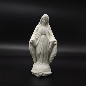 Könnte beinhalten: Eine weiße Keramikstatue einer Frau mit ausgestreckten Armen, die ein fließendes Gewand trägt. Die Statue hat einen ruhigen Ausdruck und ist vor einem schwarzen Hintergrund platziert. Der Sockel der Statue hat ein herzförmiges Design.