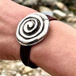 Può includere: Un braccialetto d'argento con un ciondolo a spirale. Il braccialetto è fatto di cordoncino in pelle marrone.