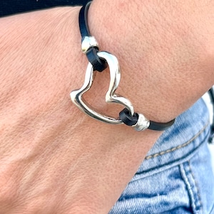 May include: A black leather bracelet with a silver heart charm. The heart charm is open and has a smooth, polished finish.