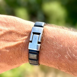May include: A silver and black bracelet with a rectangular clasp. The bracelet is made of a dark material with a silver-colored metal clasp. The bracelet is worn on a wrist.