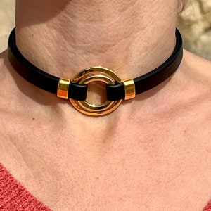 Puede incluir: Un collar de cuero negro con un colgante circular dorado. El colgante tiene un diseño de doble anillo.