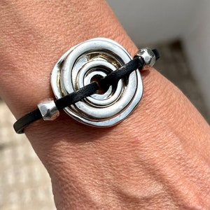 May include: A silver spiral charm bracelet with a black leather strap. The charm is a large, circular design with multiple concentric circles.