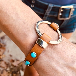 Puede incluir: Un brazalete de cuero marrón con un círculo plateado y piedras turquesa.