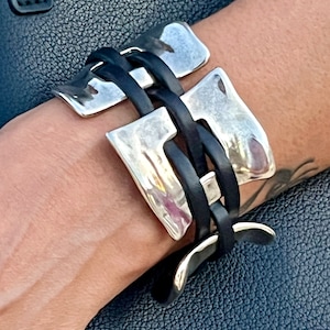 Könnte beinhalten: Ein silbernes und schwarzes Lederarmband mit einem einzigartigen, geometrischen Design. Das Armband besteht aus einer Reihe von ineinandergreifenden Silberplatten und schwarzen Lederriemen.