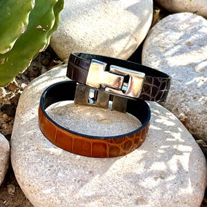 Puede incluir: Dos pulseras de cuero con textura de cocodrilo. Una es marrón, la otra de un tono más claro. Ambas tienen cierres plateados. Las pulseras se exhiben sobre piedras claras. Circunferencia aproximada de 20 cm.