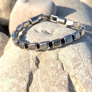 May include: A silver bracelet with square-shaped links, some of which have a black leather inlay. The bracelet is on a background of white stones.