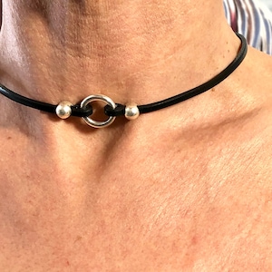 Puede incluir: Un collar gargantilla de cuero negro con un anillo de plata y dos cuentas de plata.