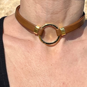 Könnte beinhalten: Ein braunes Lederhalsband mit einem goldenen Metallring. Das Halsband ist verstellbar und hat ein schlichtes Design.