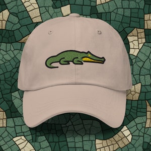 Può includere: Un cappello da baseball beige con un design di alligatore verde ricamato. L'alligatore ha la bocca e gli occhi gialli.