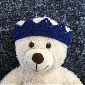 May include: A white teddy bear wearing a blue crocheted crown. The bear has a brown nose and black eyes.