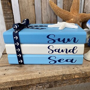 May include: A stack of three blue and white painted books with the words "Sun Sand Sea" in navy blue lettering. The books are decorated with a blue ribbon with white anchors and a white starfish.