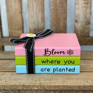 May include: Three stacked books with a black ribbon and a white daisy. The top book is pink and says "Bloom*" in black. The middle book is green and says "where you" in black. The bottom book is blue and says "are planted" in black.