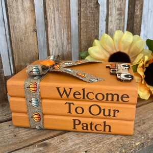 May include: A stack of three orange painted books with a burlap ribbon tied around them. The ribbon has a pattern of white and orange pumpkins. The top book has a wooden cutout of a truck with pumpkins on it. The books are stacked on a wooden surface and the text "Welcome To Our Patch" is printed on the books.