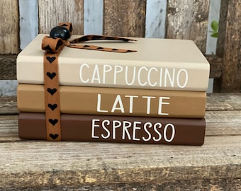 May include: Three decorative books painted in beige, tan, and brown, stacked together. The books are labelled "CAPPUCCINO", "LATTE", and "ESPRESSO" in white lettering. A brown ribbon with black hearts and a black bead is tied around the books.