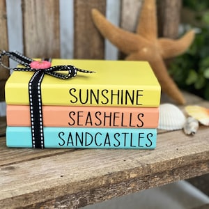 May include: Three stacked books with a summer theme. The top book is yellow and says "Sunshine", the middle book is peach and says "Seashells", and the bottom book is light blue and says "Sandcastles".