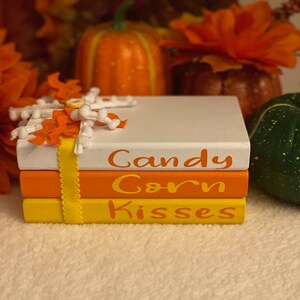 May include: A stack of three white, orange, and yellow painted books with the words "Candy Corn Kisses" written in orange on the front. The books are decorated with a white and orange fabric flower and a yellow ribbon.