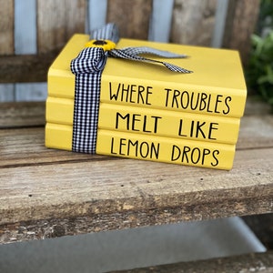 May include: Three yellow stacked books with black text that reads "Where troubles melt like lemon drops". The books are tied together with a black and white checkered ribbon and a small yellow flower.